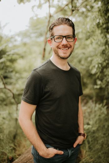 an approachable looking brown-haired man with black glasses and hands in his pockets